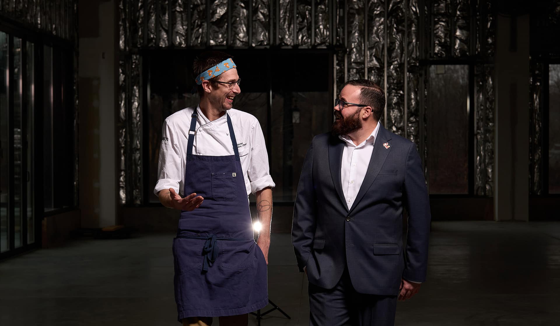 Chef Tyler and Robert van Thiel walking through a moody interior at Nowa, one in a chef's apron and one in a suit