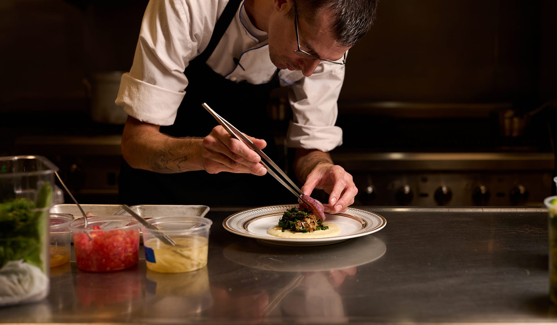 Chef Tyler leaning in to plate a dish with tweezers in low light at Nowa
