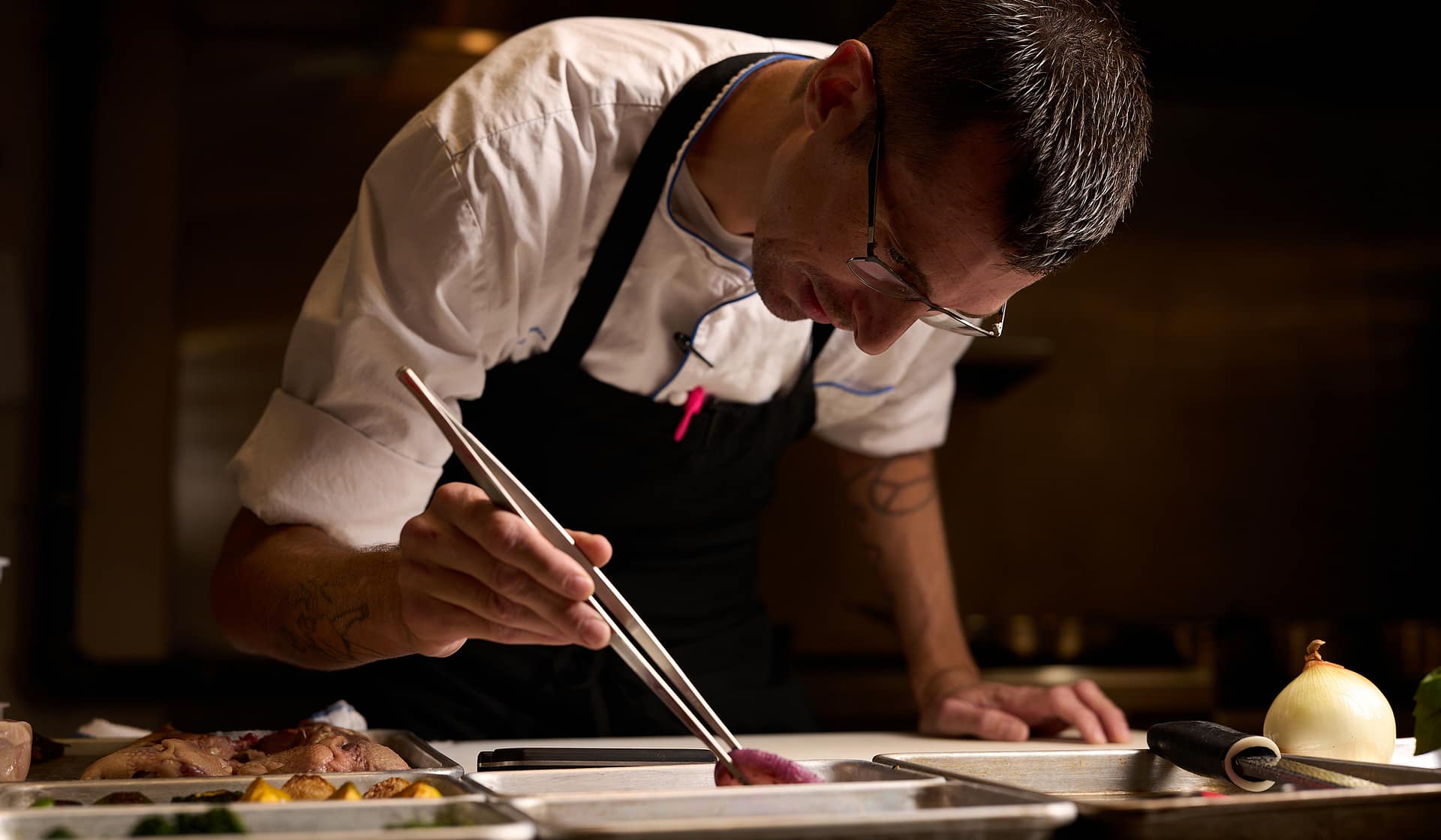 Chef Tyler plating with precision tweezers at the open kitchen counter at Nowa